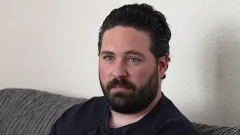 A man with slicked back black hair and black facial hair, wearing a black t-shirt while sat on a sofa