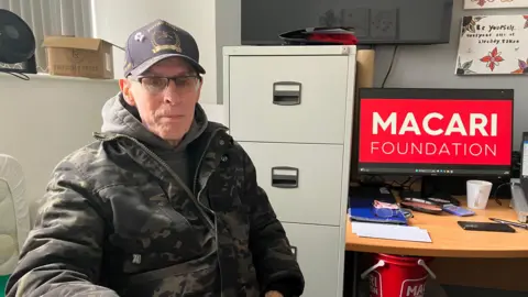 A man wearing a baseball cap and camouflage- patterned coat is sitting at a desk. Behind him is a metal filing cabinet. There is a computer with the logo of the Macari Foundation in red on it. There are buckets and boxes under another desk behind him.
