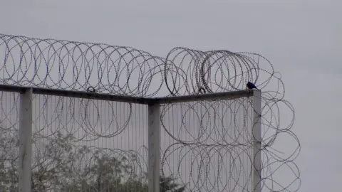 A barbed wire fence at Swinfen Hall prison. It is set against a backdrop of a grey sky. There are some trees in the background.