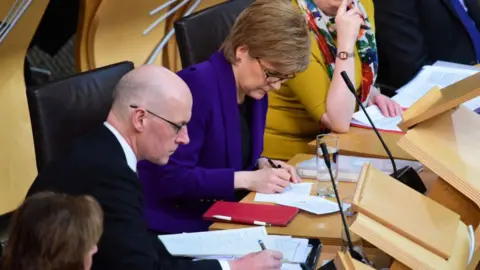 Getty Images John Swinney and Nicola Sturgeon in the Scottish Parliament chamber - both are writing on scraps of paper with pencils, with various notes strewn around the wooden desks in front of them