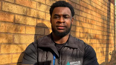 Jasheem Benson A man with black hair is wearing a t-shirt and a black gillet. He is standing in front of a brick wall with his arms crossed.