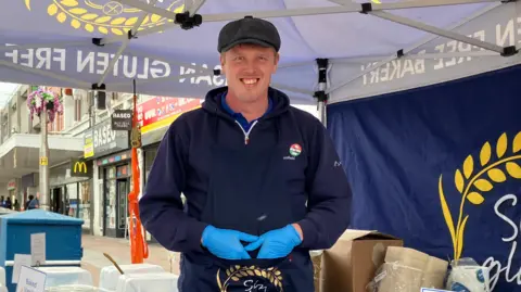 Henry Godfrey-Evans/ BBC Tom standing in a market stall, under a gazebo. He is wearing a flat cap and has blue plastic gloves on his hands. He is in Southend high street.