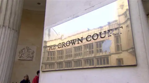 A brass plaque on a sand stone wall reading The Crown Court. A coat of arms is visible on a wall in the background. 