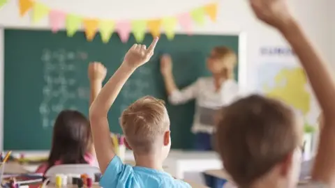 Getty Images File picture of a classroom. Three children can be seen, from behind, with their hands up. One is wearing a blue T-shirt. A teacher is at the blackboard in the background, with her right hand pointing to some writing on the board. There is a map on the wall to the right.