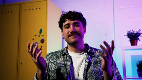 A young man with a moustache, dark shirt and white T-shirt.