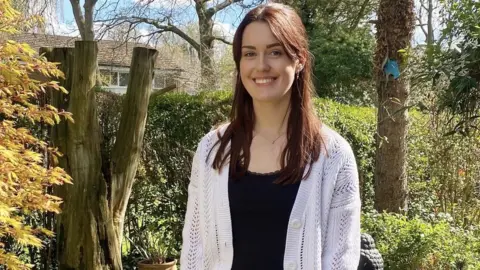 Emily Katy  Emily has long brown hair, tied back in a half-up, half down style with bangs. She's wearing a cream cardigan and black vest top. She's standing in a garden. You can see autumnal trees, hedges and the top of a house in the background.