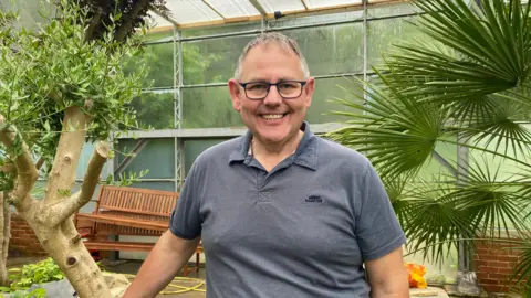 A middle-aged man with short grey hair and glasses. He is wearing a grey polo top and smiling as he looks into the camera. He is stood in a large green house with green plants behind him
