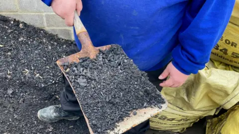 A close-up of a hand shovel full of black ash with yellow bags in the background.