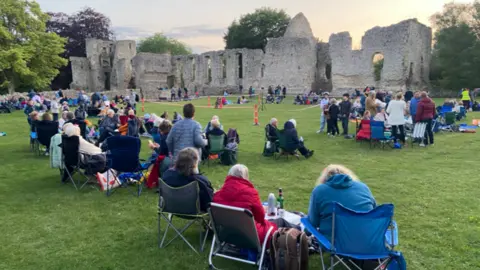 A ruined stone building surrounded by scores of people sitting in picnic chairs.