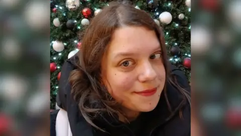 Natasha Alsop stands in front of a Christmas tree with black, red and white baubles and fairy lights on it. She has short brown hair and wears a back puffer coat.
