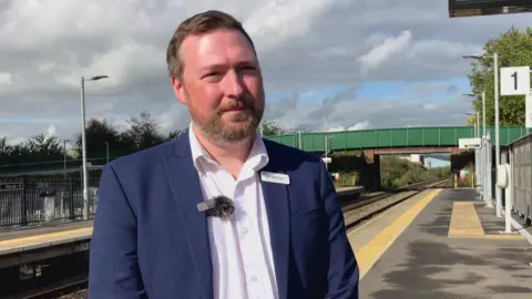 A man with a dark hair, a bread, wearing a blue blazer, stood on a railway platform 