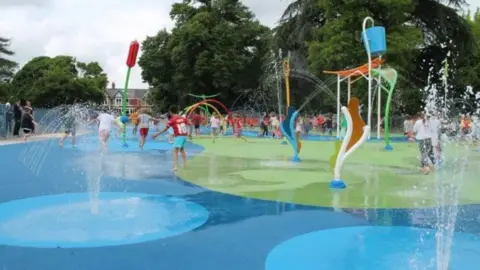 Kids playing at a splash park with a number of water features spraying water in the area. There are parents stood around the perimeter of the splash area watching with trees seen in the background.