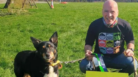 The picture shows a man and his border collie dog that is attached to a leesh. The man is kneeling down, close to his dog on bright green grass. Behind him is a cone and a wooden pallet
