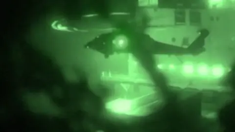 A helicopter hovers in front of a ship in footage shot at night