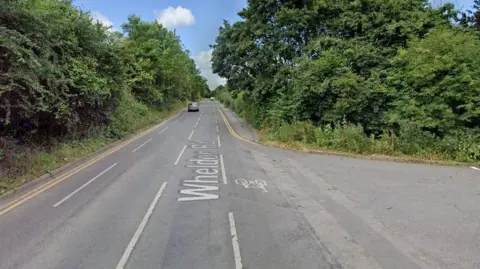 Wheldon Road, single-carriage road, pictured on a sunny day with trees and bushes at the roadside. There is a slim pavement on one side of the road and double yellow lines on both sides. 