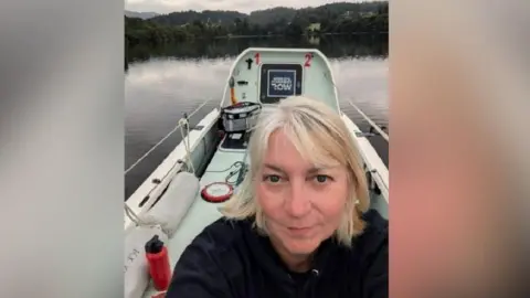 Vivienne Barclay sat on a boat on a lake with scenery behind her of dark green trees and hills. She has light hair and is wearing a dark hooded jumper. Next to her on the boar is a red water bottle with a black cap. 