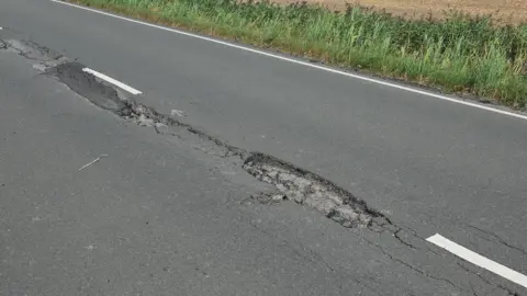 A road on tarmac with a cracked part in the middle is next to a grass verge.