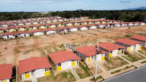 Rows of identical grey and yellow houses with red roofs lining roads, with plots of empty ground behind each house and forest-covered hills in the background, in Isberyala