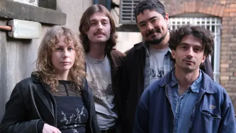 Four members of Brown Horse standing together and looking at the camera outdoors in front of brick and concrete walls.