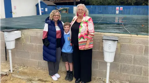 Leonard Stanley Primary School's Office manager Lisa Williams stands in front of the pool with head of the Parent Teacher Association Ally Higgins and her son who is a pupil at the school. The pool is still under construction and has a covering on it.