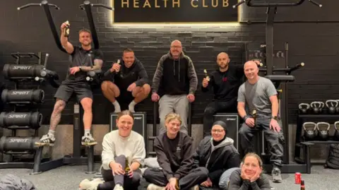Josh Baines A group of people sat surrounded by gym equipment and in front of a sign which says 'The Atrium Health Club'
