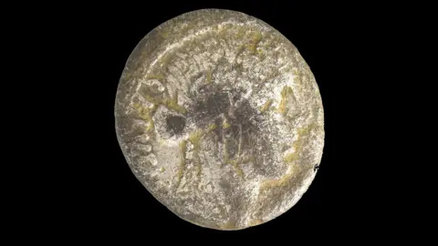 Friends of the Oxfordshire Museum A round Roman coin - it is not perfectly round but has uneven edges. A pattern can be seen on its surface.