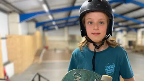 Bobby is standing on a ramp in a warehouse which is a skatepark. He is wearing a black helmet and blue T-shirt and is holding a skateboard, which has stickers on it, across his body. Bobby has shoulder length blonde hair.