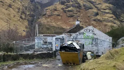 A vandalised one-storey white house with no roof is surrounded by a wire fence. There is a full skip out the front. The house sits in a scenic area with hills in the background.