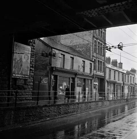 Bert Hardy/Getty Images A man walking under a railway bridge and past a row of terraced shops