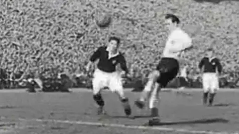 A black‑and‑white action shot shows two footballers contesting a bouncing ball near the penalty area. One player is lifting a leg to strike while another moves in to challenge, with additional players further back. A tightly packed crowd fills the stadium stands in the background, highlighting the scale and atmosphere of the match.