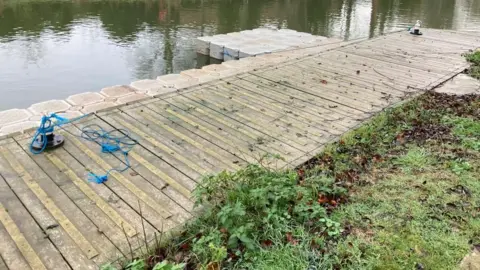 Alex Pope/BBC A wooden slatted landing stage, by a river bed, with blue rope round a metal clip, grass and plants in front and water to the left. Plastic pontoons are in front of it. 