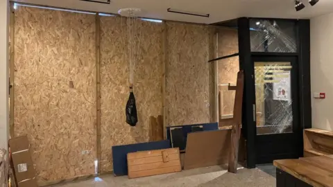 The inside of a boarded up shopfront with four panels. Grey wires hang from the ceiling and are tied up by a black plastic bag. On the right there is a shattered shop door.