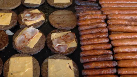 A griddle filled with cheese and bacon topped burgers and sausages