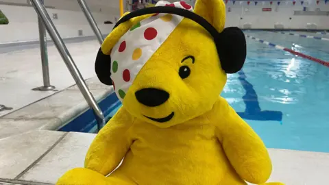 A Pudsey plush toy with ear muffs sat on the side of a swimming pool