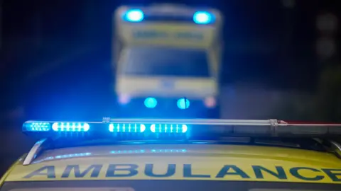 A close-up on the top of an ambulance is at the bottom of the photo, showing the word ambulance and blue flashing lights. The background is blurred but another ambulance is behind it.