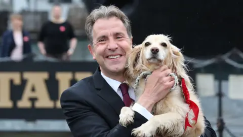 Metro Mayor and MP Dan Norris smiles at the camera while holding up his golden spaniel dog who wears a red rosette 