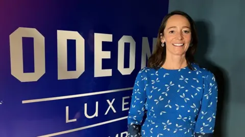 James Grant/BBC A woman in a blue dress, smiling and looking at the camera, next to a Odeon LUXE sign.
