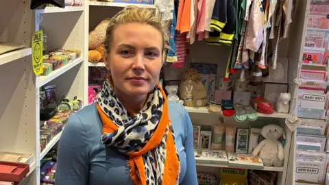 Sophie Bryant has blonde hair tied back and is wearing a blue long-sleeved top and a leopard print scarf with an orange trim.