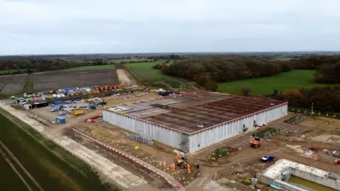 John Fairhall/BBC A drone image of a development site. A large square building is in the process of being built. Workers in orange hi-vis suits can be seen working around the site. Other building infrastructure can seen around the square building.