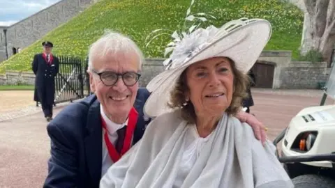 Geoff Almond has write hair, black rimmed glasses, a blue suit, white shirt with black time and a red lanyard round his neck. Jean Almond has a white broads brimmed hat with floral decorations a white blouse and grey shawl.