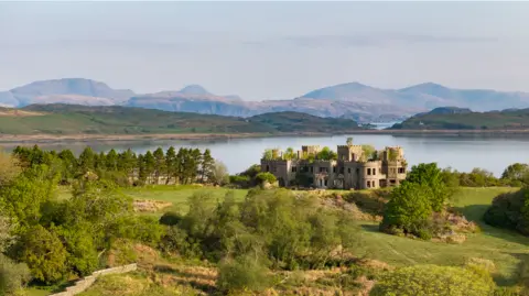 The ruins of Shuna Castle, a flat-roofed turreted castle, are surrounded by grassy banks and trees and overlooking Loch Melfort with a view in the distance of mountains.