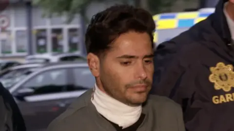 Ali Sohrabi, a man with short, black hair and short stubbly beard, being escorted to Galway District Court by gardaí (Irish police). He is wearing a grey or khaki green top and there is a white scarf or possibly a bandage around his neck. The garda officer beside him is wearing a blue uniform jacket with a gold garda crest.