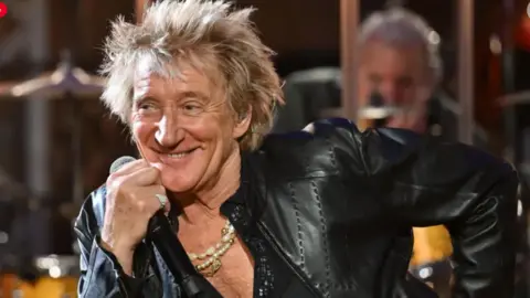 A close up of Rod Stewart in a leather jacket and holding a microphone, smiling at someone to the right of camera