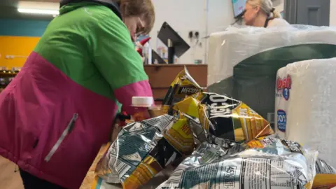 A woman wearing a pink and green jacket loads shopping on to a counter at a community grocery shop, while an assistant looks on in the background.