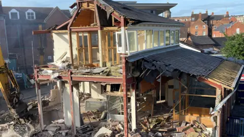 A closer shot of the building that has been half-demolished. You can see the inside of building, and there is wood, brick and rubble on the floor