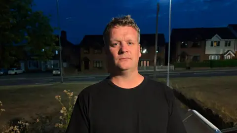 A man in a black t-shirt with grey hair, he is looking towards the camera with a stern look.