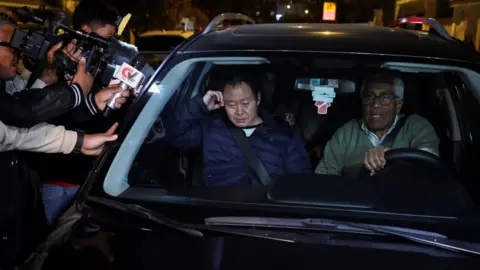 Reuters Kenji Fujimori, son of Peruvian former President Alberto Fujimori, leaves Keiko Fujimori's house in Lima. He is sat in the passenger seat of a vehicle with TV camera crews and reporter's microphones seen in the shot as he is driven away. Kenji is visibly upset.