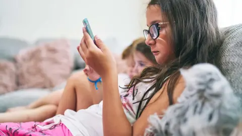 A anonymous young girl looking at her phone on her bed in a room with brightly coloured cuddly toys. She is wearing a pink flowery jumper and jeans, we cannot see her face.
