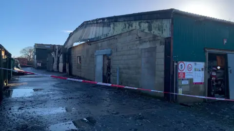 Martin Allen Red and white tape stretches across in front of a concrete block-built building with corrugated iron at the top which is scorched. Further buildings stand next to it, one with buckled metal walls. The concrete ground in front of them has several puddles on it.