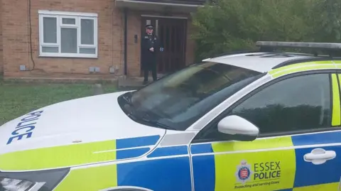 A white, yellow and blue marked fluorescent police car is parked outside an address in Brentwood. The car is stationary and outside the front door of the property stands a police officer wearing police uniform.